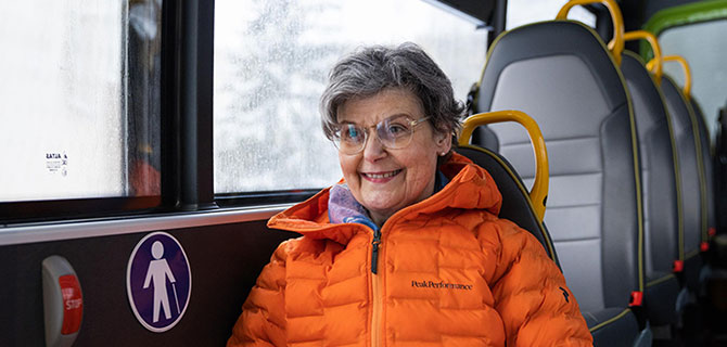 Smilende kvinne i oransje jakke sitter om bord i en minibuss. Utenfor er det vått og tåkete.