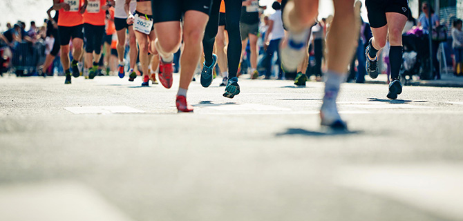 A group of runners participating in a marathon. The focus is on their legs and feet as they run along a city street, wearing various running shoes and athletic gear.