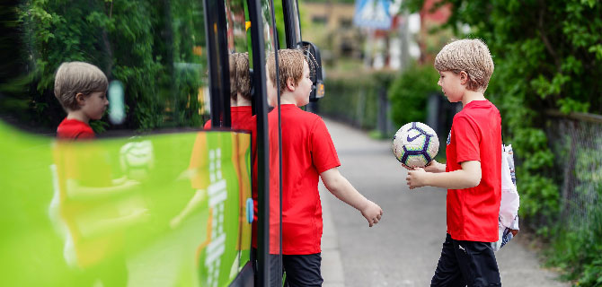 To gutter i røde t-skjorter i ferd med å gå om bord i en grønn buss. Den ene holder en fotball, og speilbildet hans vises i bussens vindu. Bakgrunnen er grønn og frodig.
