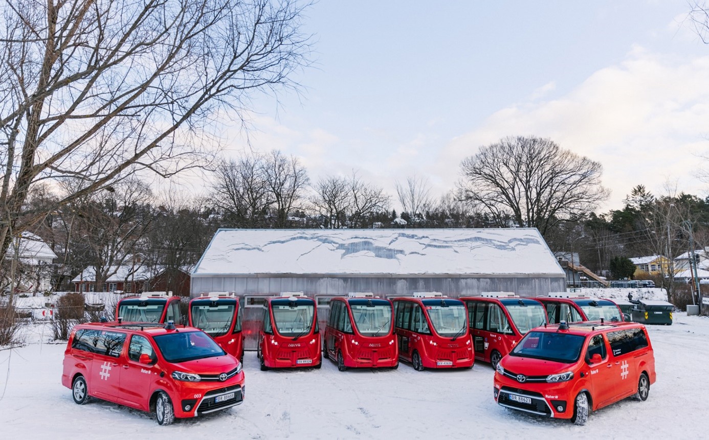 Ni røde selvkjørende kjøretøy parkert en vinterdag. Et skur og flere trær i bakgrunnen.